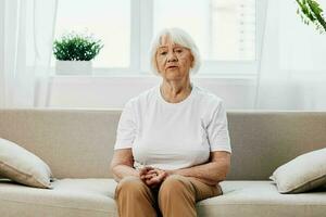 Elderly woman sits on sofa at home, bright spacious interior in old age smile, lifestyle. Grandmother with gray hair in a white T-shirt and beige trousers. photo