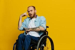 A man in a wheelchair problems with the musculoskeletal system looks at the camera is not satisfied, with tattoos on his arms sits on a yellow studio background, health concept man with disabilities photo