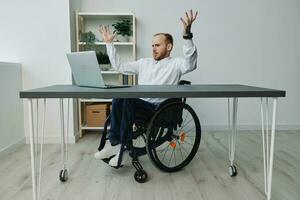 un hombre en un silla de ruedas empresario con tatuajes en el oficina detrás un computadora portátil, ira y molestia, manos arriba, el concepto de trabajando un persona con discapacidades, libertad desde social marcos foto