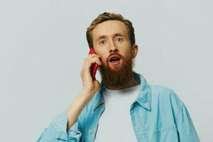 Man hipster with phone in hand blogger smile on gray background in blue shirt and white t-shirt, talking on the phone and selfies photo