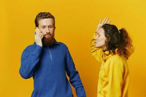 Man and woman couple with phone in hand call talking on the phone, on a yellow background, symbols signs and hand gestures, family quarrel jealousy and scandal. photo