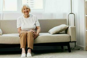 elderly woman sits on a sofa at home against the backdrop of a window and a happiness smile, stylish interior. Lifestyle retirement. photo