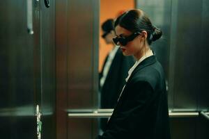 Stylish woman in black jacket and sunglasses posing in elevator, fashion model, dark cinematic light and color, glamor vintage photo