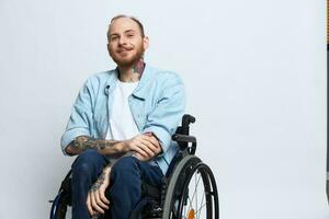 un hombre en un silla de ruedas sonrisa, Copiar espacio, con tatuajes en su brazos se sienta en un gris estudio fondo, el concepto de salud es un persona con discapacidades, un real persona foto