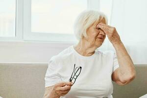 Elderly woman severe eye pain sitting on the couch, health problems in old age, poor quality of life. A grandmother with gray hair holds on to her sore eyes with glasses in her hand. photo