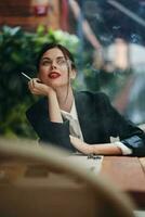 elegante Moda mujer retrato sentado en un café a un mesa y de fumar un cigarrillo liberando fumar desde su boca con rojo lápiz labial, un malo hábito, un sonrisa con dientes y un pensativo Mira foto