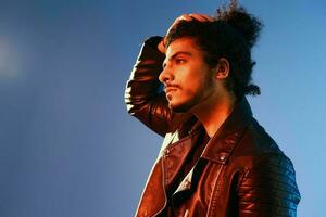 Portrait of a stylish man with curly hair on a blue background multinational, colored light, black leather jacket trend, modern concept. photo