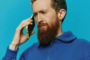 retrato de un hombre con un teléfono en su manos lo hace mirando a eso y hablando en el teléfono, en un azul antecedentes. comunicado en línea social medios de comunicación, estilo de vida foto