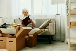 elderly woman sits on a sofa at home with boxes. collecting things with memories albums with photos and photo frames moving to a new place cleaning things and a happy smile.