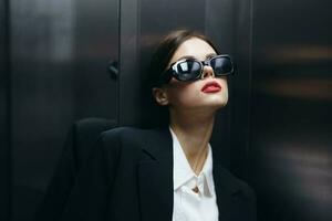Stylish woman in a black jacket and sunglasses posing in an elevator lift near the mirror, fashion model, dark cinematic light and color matrix, glamor vintage photo