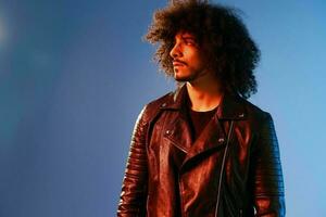 Portrait of a stylish man with curly hair on a blue background multinational, colored light, black leather jacket trend, modern concept. photo