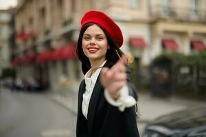 Moda mujer sonrisa con dientes en pie en el calle en frente de el ciudad turista seguir yo elegante ropa con rojo labios y rojo boina, viajar, cinematográfico color, retro Clásico estilo, urbano moda. foto