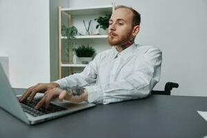 un hombre en un silla de ruedas un empresario con tatuajes en el oficina trabajos a un computadora portátil, integración dentro sociedad, el concepto de trabajando un persona con discapacidades, libertad desde social marcos foto