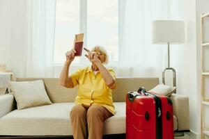 Happy senior woman with passport and travel ticket packed a red suitcase, vacation and health care. Smiling old woman joyfully sitting on the sofa before the trip raised her hands up in joy. photo