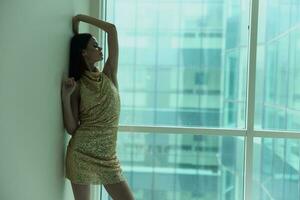 Woman in gold sequined dress by the window poses in a stylish image designer dress with open back, party look photo