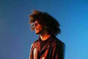 Portrait of a stylish man with curly hair with glasses and headphones on a blue background multinational, colored light, black leather jacket trend, modern concept. photo