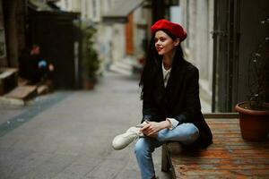 Stylish woman tourist in stylish clothes in a jacket and red beret sitting outside with phone in hand, cell phone in travel, French style, cinematic color. photo