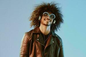 Portrait of a stylish man with curly hair with glasses and headphones on a blue background multinational, colored light, black leather jacket trend, modern concept. photo