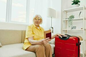 Happy senior woman with passport and travel ticket packed a red suitcase, vacation and health care. Smiling old woman joyfully sitting on the sofa before the trip raised her hands up in joy. photo