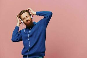 Portrait of a redheaded man wearing headphones smiling and dancing, listening to music on a pink background. A hipster with a beard. photo