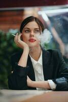 elegante mujer se sienta en un café a un mesa y fuma un cigarrillo liberando fumar desde su boca, un malo hábito, un sonrisa con dientes y un pensativo Mira foto