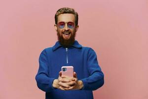 Portrait of a redheaded man with phone in hand taking selfies and photos on his phone with a smile on a pink background, blogger