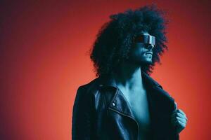 Portrait of fashion man with curly hair on red background with stylish glasses, multicultural, colored light, black leather jacket trend, modern concept. photo