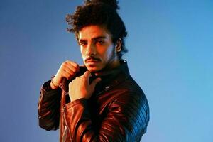 Portrait of a stylish man with curly hair on a blue background multinational, colored light, black leather jacket trend, modern concept. photo