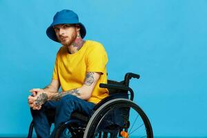 A man in a wheelchair problems with the musculoskeletal system looks at the camera in a t-shirt with tattoos on his arms sits on a blue studio background, full life, real person, health concept photo