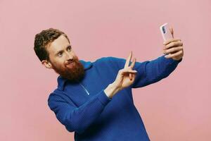 Portrait of a redheaded man with phone in hand taking selfies and photos on his phone with a smile on a pink background, blogger