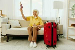 elderly woman with passport and travel ticket packed a red suitcase, vacation and health care. Smiling old woman joyfully sitting on the sofa before the trip raised her hands up the joy of retirement. photo