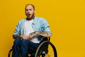 un hombre en un silla de ruedas sorprendido, con tatuajes en su brazos se sienta en un amarillo estudio fondo, el concepto de salud es un persona con discapacidades foto