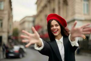 Moda mujer sonrisa con dientes en pie en el calle en frente de el ciudad turista seguir yo elegante ropa con rojo labios y rojo boina, viajar, cinematográfico color, retro Clásico estilo, urbano moda. foto