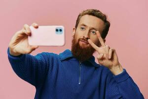 Portrait of a redheaded man with phone in hand taking selfies and photos on his phone with a smile on a pink background, blogger