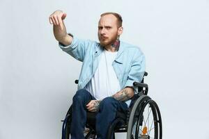 A man in a wheelchair and pointing a finger at, copy space, with tattoos on his hands sits on a gray studio background, health concept a person with disabilities, a real person photo