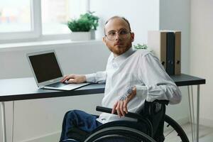 un hombre en un silla de ruedas mirando a el cámara empresario en el oficina trabajando en un ordenador portátil en línea, social redes y puesta en marcha, integración dentro sociedad, trabajo concepto hombre con discapacidades foto