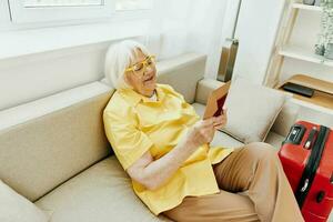 Happy senior woman with passport and travel ticket packed a red suitcase, vacation and health care. Smiling old woman joyfully sitting on the sofa before the trip raised her hands up in joy. photo