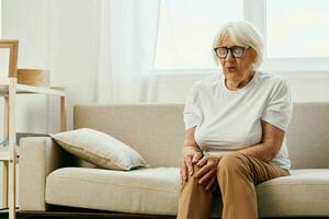 Elderly woman severe pain in her leg sitting on the couch, health problems in old age, poor quality of life. Grandmother with gray hair holds on to her sore knee, problems with joints and ligaments. photo