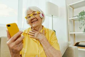 Happy elderly woman watching video call on phone and listening to music on headphones, surprise and open mouth, bright modern interior, lifestyle online communication. photo