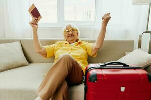 Happy senior woman with passport and travel ticket packed a red suitcase, vacation and health care. Smiling old woman joyfully sitting on the sofa before the trip raised her hands up in joy. photo