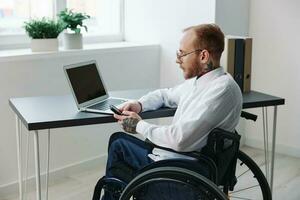 A man wheelchair in the office works at a laptop with a phone, working online, social networks and startup, integration into society, the concept working person with disabilities, real person close-up photo