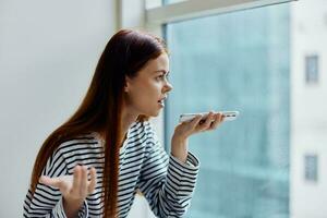 Woman holding phone and talking on video call recording via messenger voice message, view of city through windows photo