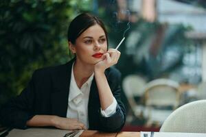 elegante Moda mujer retrato sentado en un café a un mesa y de fumar un cigarrillo liberando fumar desde su boca con rojo lápiz labial, malo hábito, sonrisa con dientes y pensativo mirar, cinematográfico Clásico foto