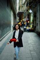 Moda mujer turista en elegante ropa en un chaqueta y rojo boina corriendo abajo el estrecho calle de el ciudad sonrisa y felicidad viaje, francés estilo, cinematográfico color. foto