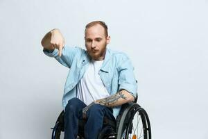 A man in a wheelchair and pointing a finger at, copy space, with tattoos on his hands sits on a gray studio background, health concept a person with disabilities, a real person photo