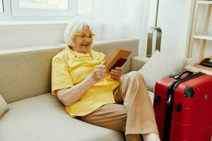 Happy senior woman with passport and travel ticket packed a red suitcase, vacation and health care. Smiling old woman happily sits on the sofa before the trip. photo