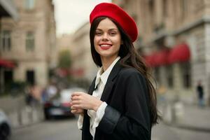 Moda mujer retrato sonrisa con dientes en pie en el calle en frente de el ciudad turista en elegante ropa con rojo labios y rojo boina, viajar, cinematográfico color, retro Clásico estilo, urbano moda. foto