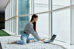 A woman sits in an office lobby by large windows and works on her laptop online via the Internet, freelance work anywhere in the world photo