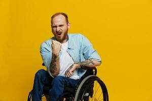man in a wheelchair screams angrily, shows fist looks at the camera is not pleased, with tattoos on his hands sits on yellow background of the studio, the concept of health is person with disabilities photo