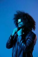 Portrait of fashion man with curly hair with stylish glasses on blue background multinational, colored light, black leather jacket trend, modern concept. photo